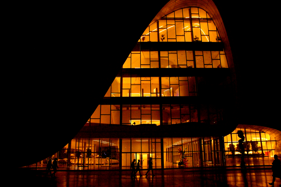 Silhouetted people walk past a glowing modern building with glass facades at night.
