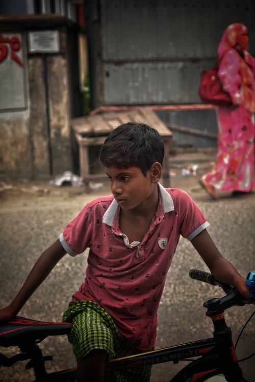 Free South Asian boy on bicycle in a bustling urban street, with a woman in traditional clothing in the background. Stock Photo