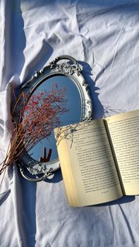 Vintage mirror and open book with dry flowers on white fabric in natural light.