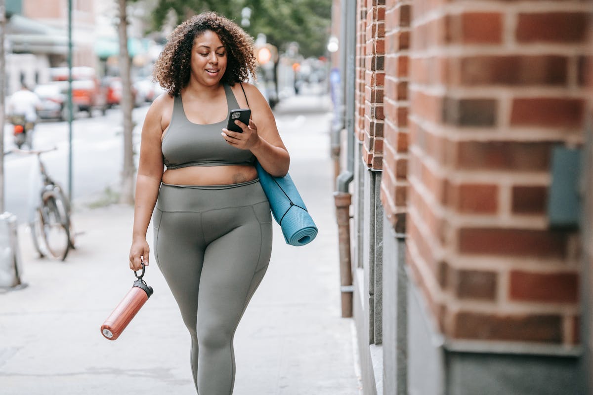 Woman Walking in Sport Clothing Carrying a Yoga Mat and Using Her Phone