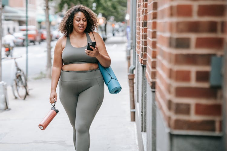 Woman Walking In Sport Clothing Carrying A Yoga Mat And Using Her Phone 