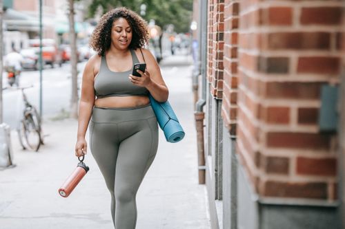 Free Confident woman in sportswear walking outside with yoga mat and phone, embracing positivity. Stock Photo