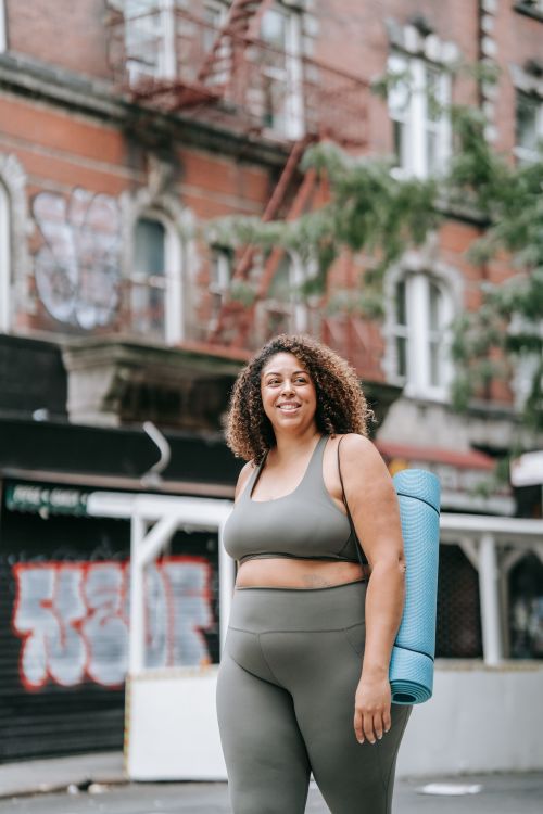Free Smiling woman in sportswear holding a yoga mat on a city street, embodying positivity. Stock Photo