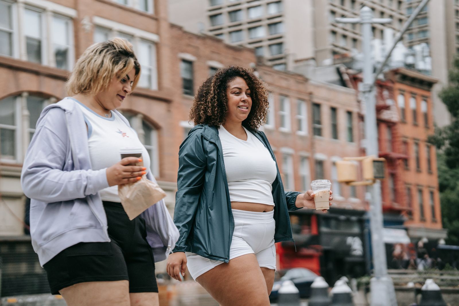Two women in casual athletic wear walking together on a city sidewalk, smiling and holding iced coffee drinks, with brick buildings lining the street behind them