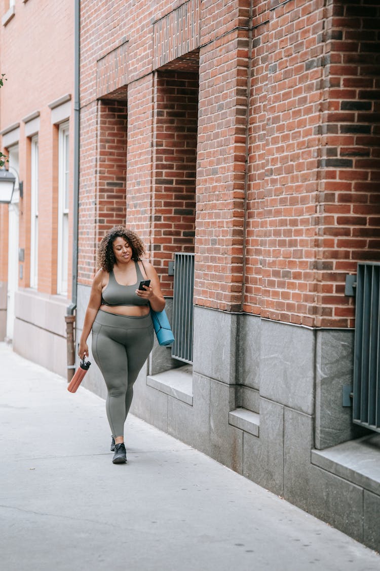 Woman Walking In Sport Clothing Carrying A Yoga Mat And Using Her Phone 