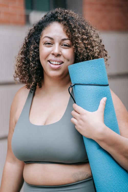 Free Confident woman smiling outdoors holding a blue yoga mat, embracing body positivity. Stock Photo