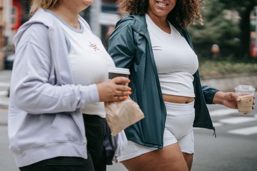 Free Two women enjoying a walk while holding coffee cups, wearing casual sports attire in a city setting. Stock Photo