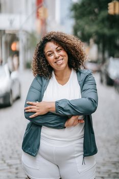 A woman with curly hair smiling confidently while standing on a cobblestone street in the city.