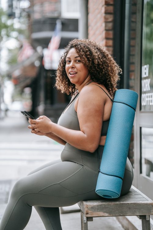 Free Smiling woman sitting outdoors with yoga mat, checking phone in sports attire. Stock Photo