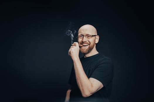 Caucasian bald man with a beard smiling and smoking against a black background in a studio setting.