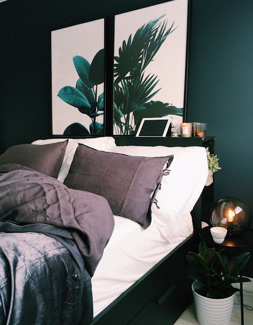 A two panel painting of green palm plants hanging over a bed with white and grey colored linens. 