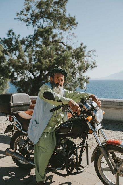 Elderly man in traditional attire with a motorbike by the sea under sunny skies.