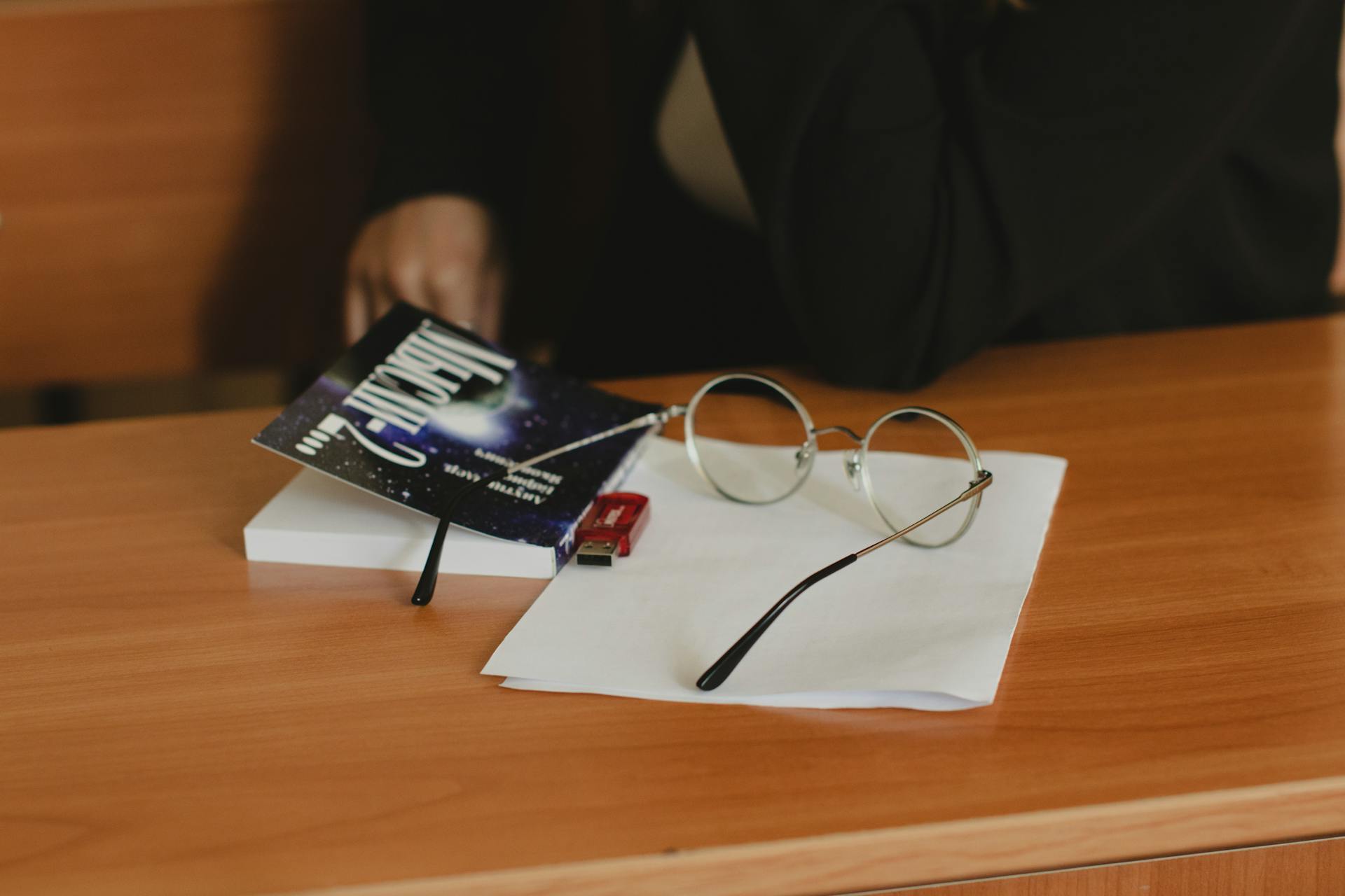 Close-up of eyeglasses, book, and USB drive on a wooden desk indoors.