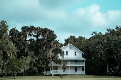 Free A quaint white house surrounded by lush trees under a blue sky, perfect serene setting. Stock Photo