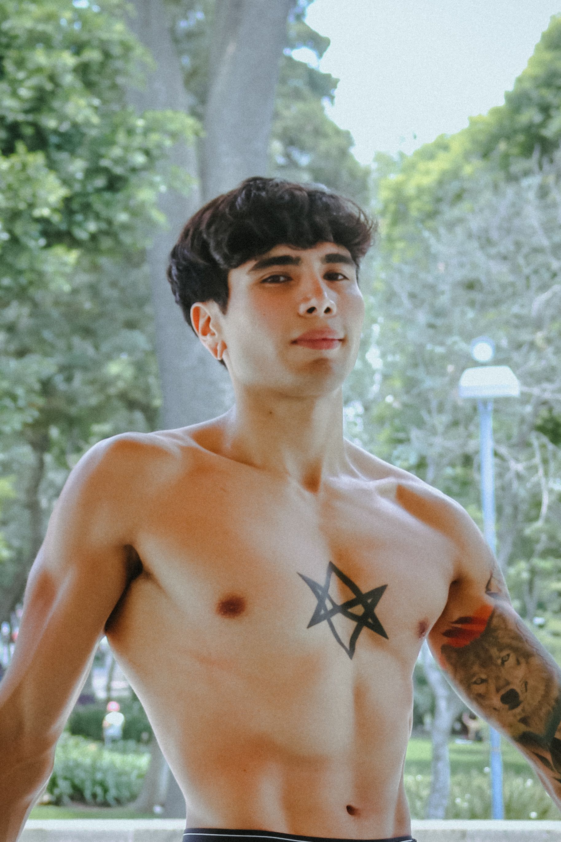 Young tattooed man poses shirtless outdoors in a park setting, showcasing confidence and style.