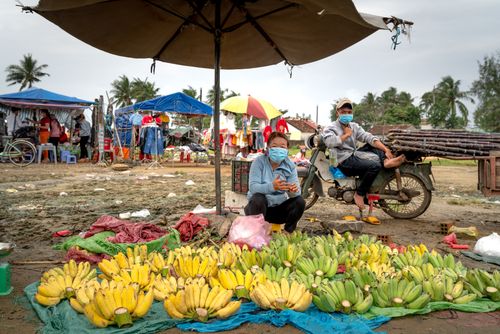 Free Two people wearing masks selling bananas at an outdoor market under an umbrella. Stock Photo