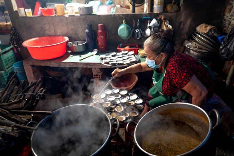 Old Woman In Face Mask Cooking In Big Pots