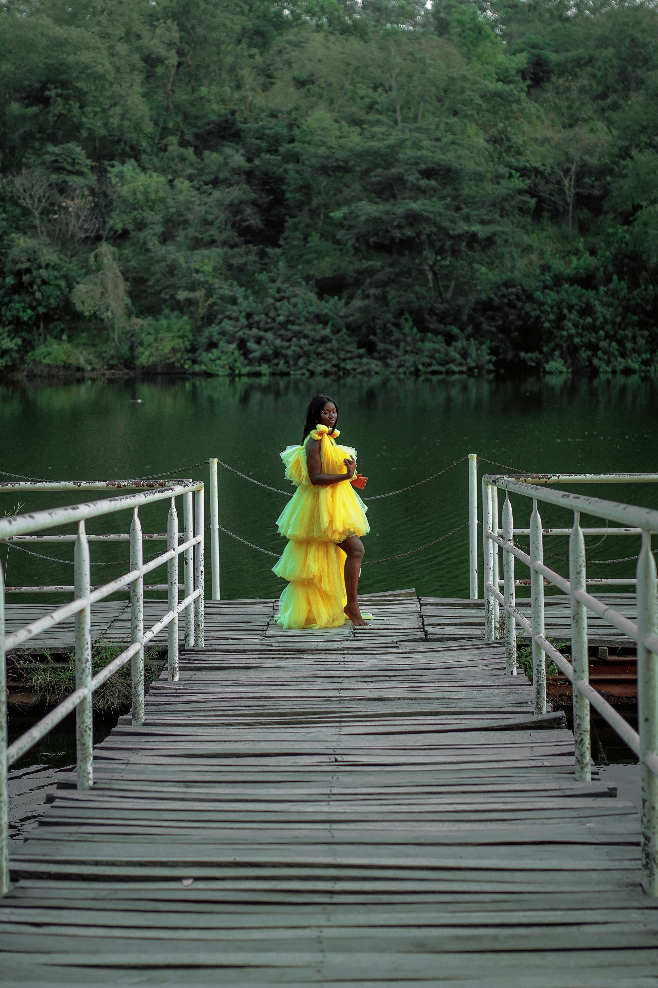 A woman in a yellow dress stands gracefully on a boardwalk by the lake.