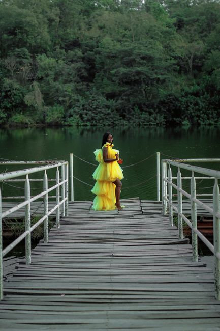 A woman in a yellow dress stands gracefully on a boardwalk by the lake.