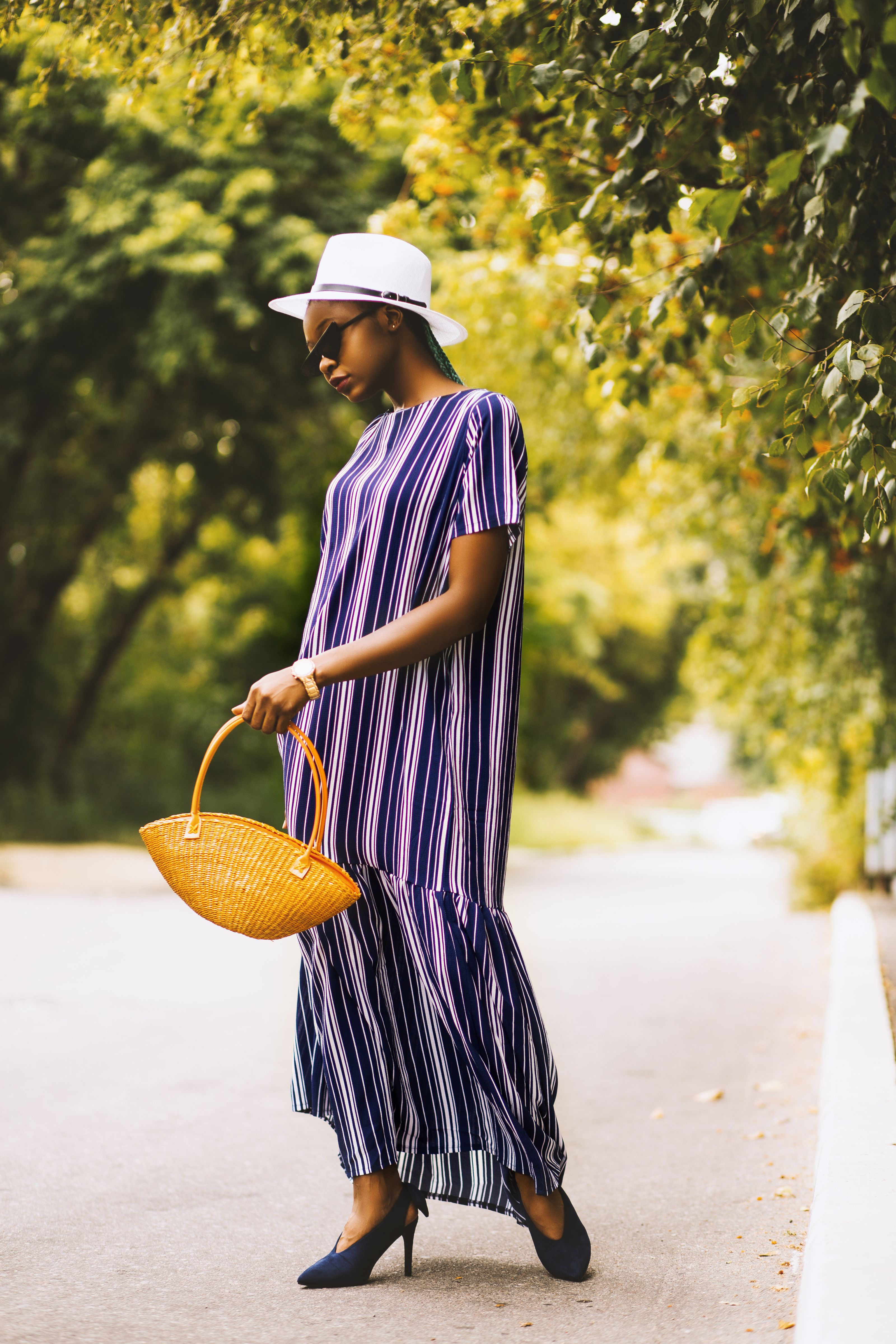 Mulher elegante em um vestido listrado e chapéu caminhando por um caminho ladeado de árvores.