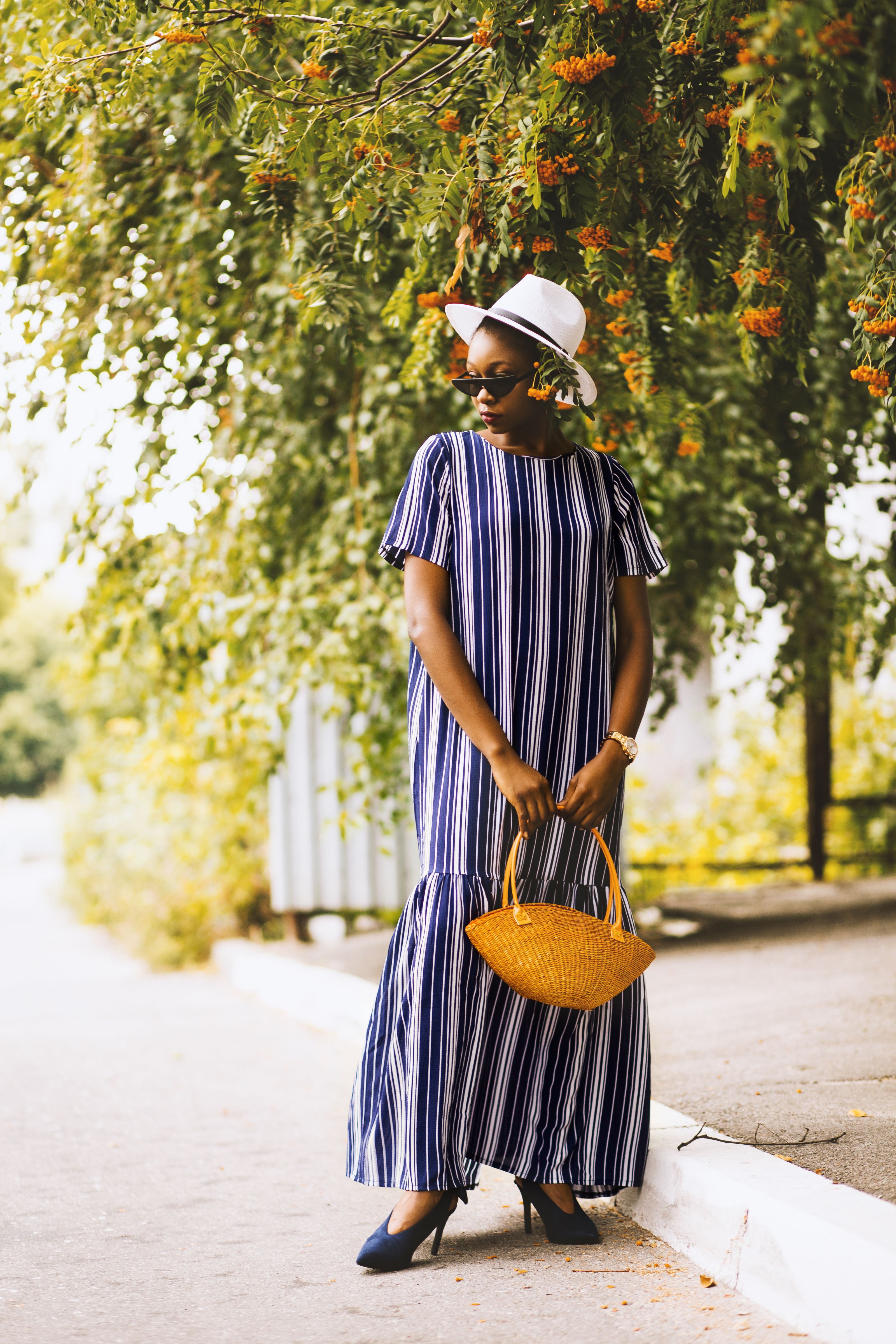 Mulher elegante em um vestido listrado e chapéu posando com uma bolsa em um dia ensolarado no parque.