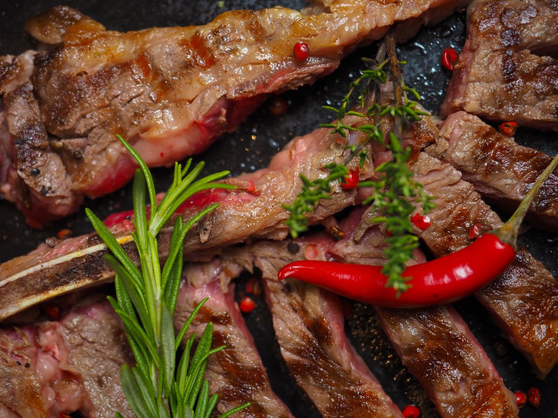 Sliced grilled bone-in steak with rosemary sprigs, red chili and pink peppercorns on dark iron