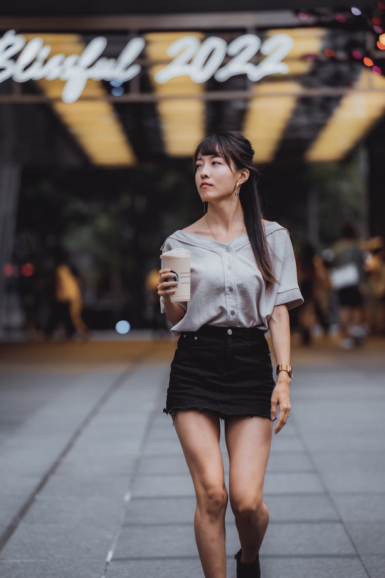 Young Woman In Gray Shirt And Black Mini Skirt Walking On Pavement Holding Coffee In A Disposable Cup