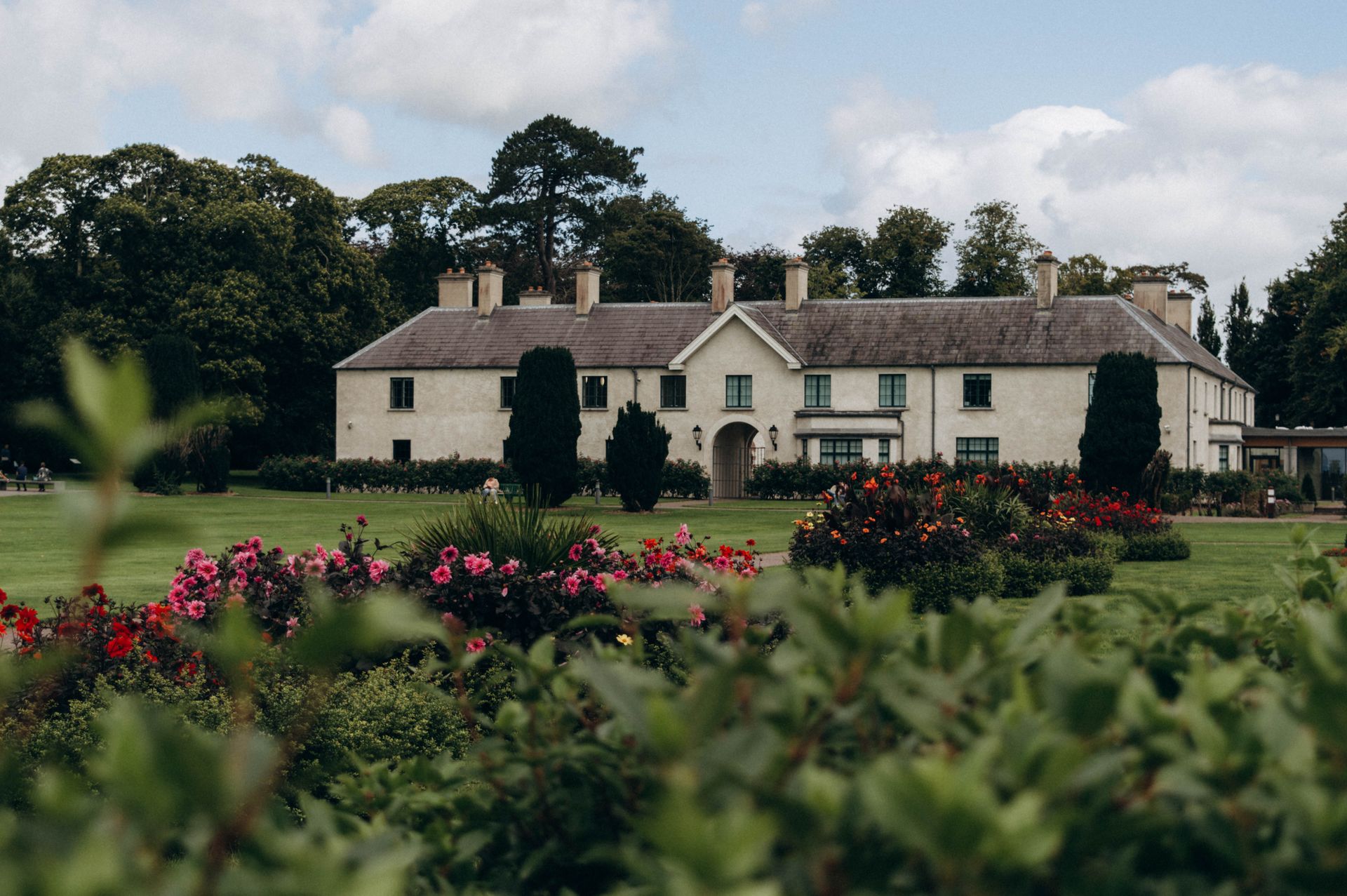 Charming manor house surrounded by vibrant flowers and lush greenery under a cloudy sky.