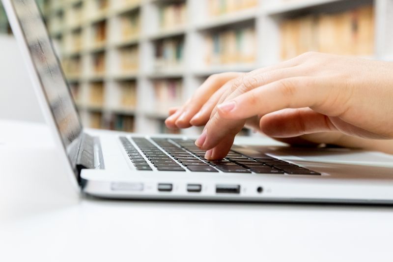 A hand typing on a laptop keyboard, with blurred bookshelves in the background