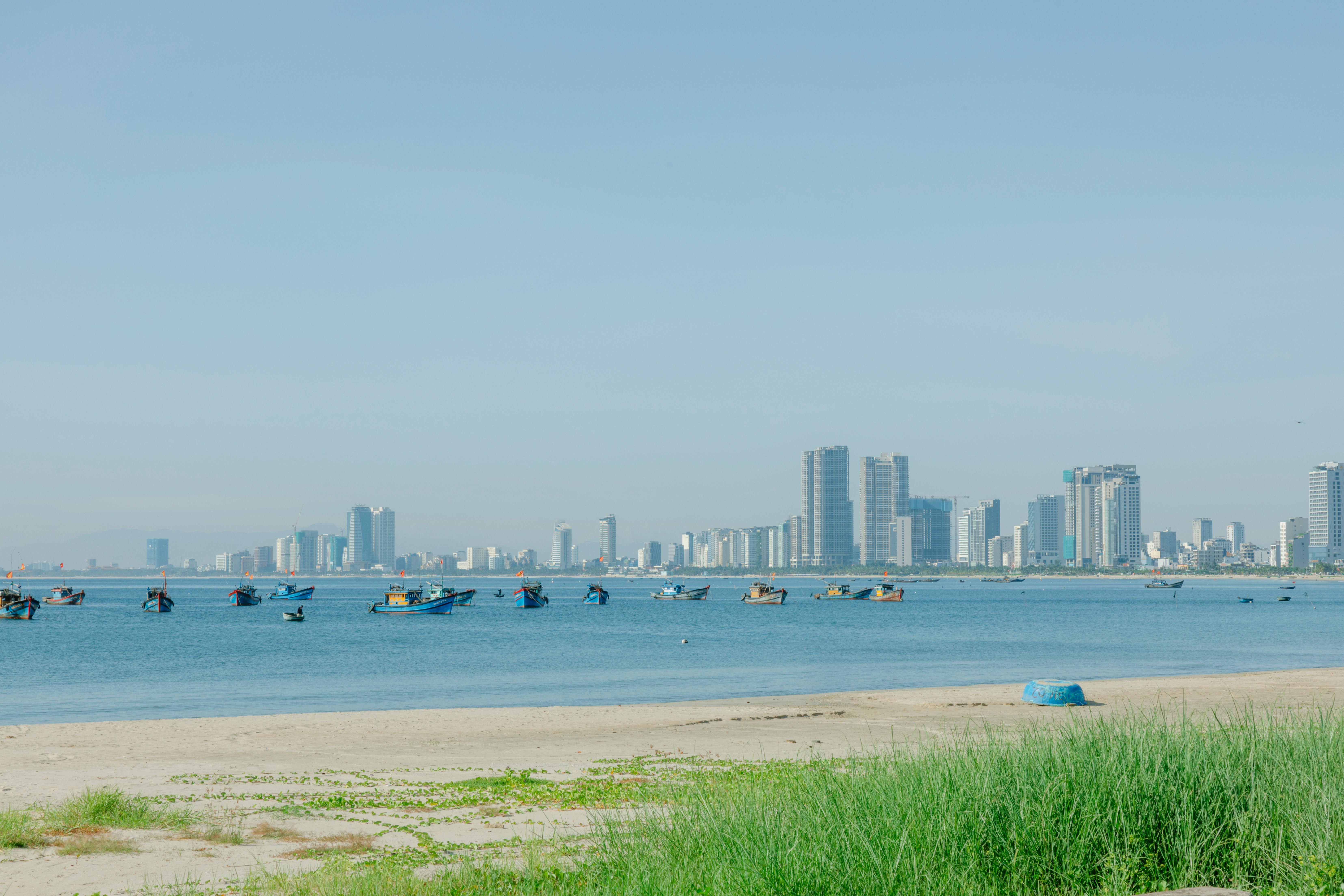 City skyline with boats on a calm ocean and sandy beach, under a clear blue sky.