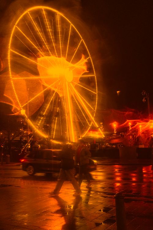 Free A vibrant ferris wheel lights up the night, creating dynamic reflections and a lively street scene. Stock Photo