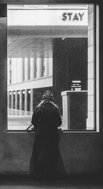 Black and white photo of person in coat and beanie looking out of window at city street.