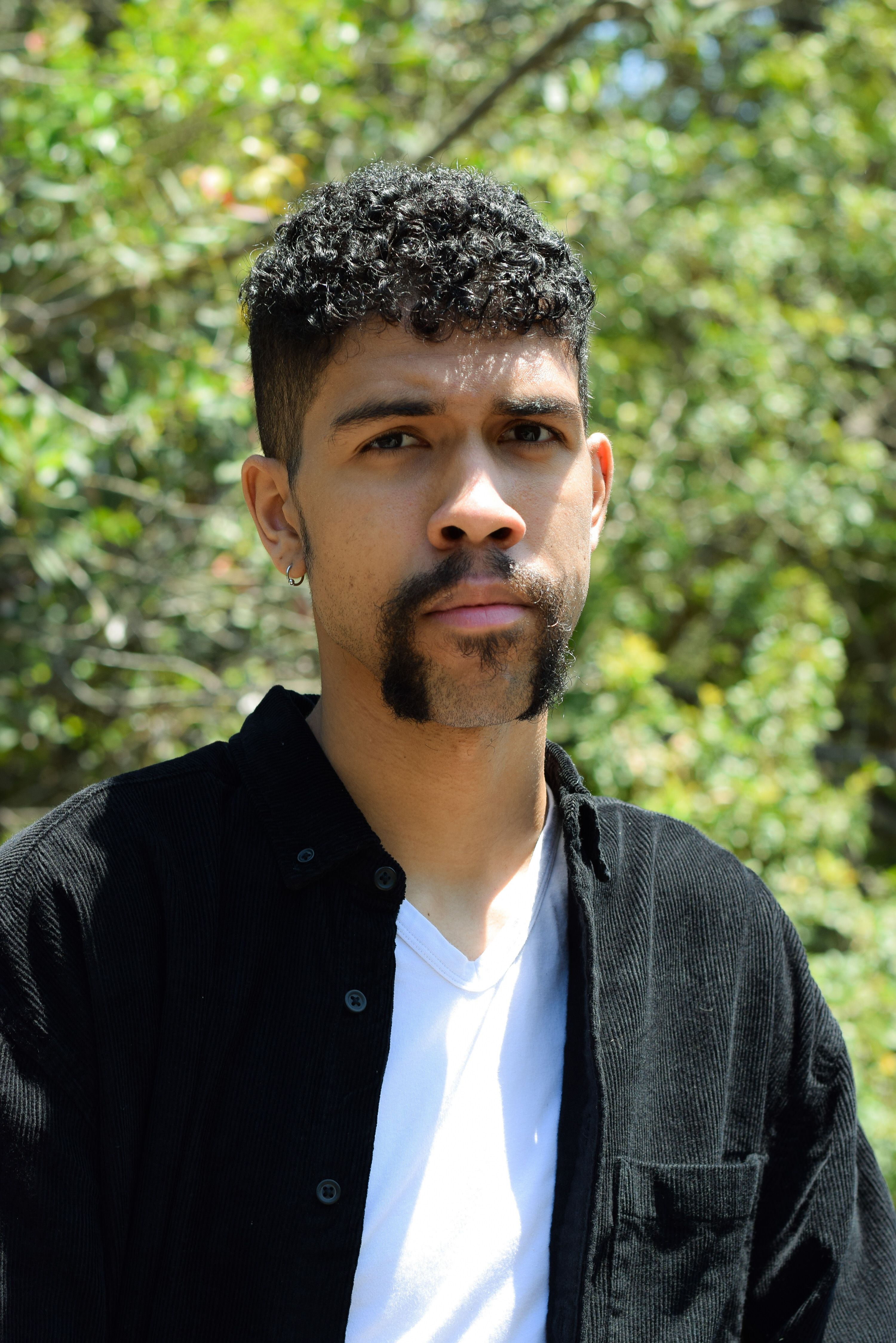 A man with facial hair poses outdoors in natural light, surrounded by greenery.