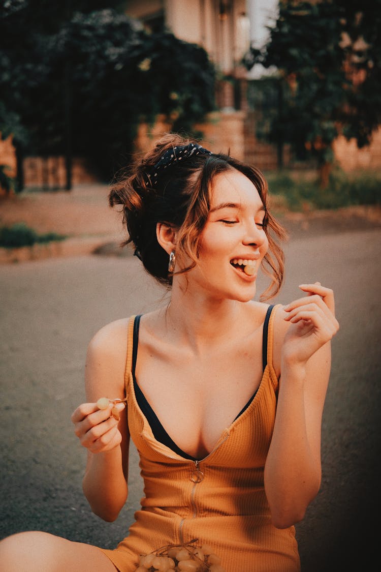 A Woman In Spaghetti Strap Top Sitting On The Ground While Eating Grapes