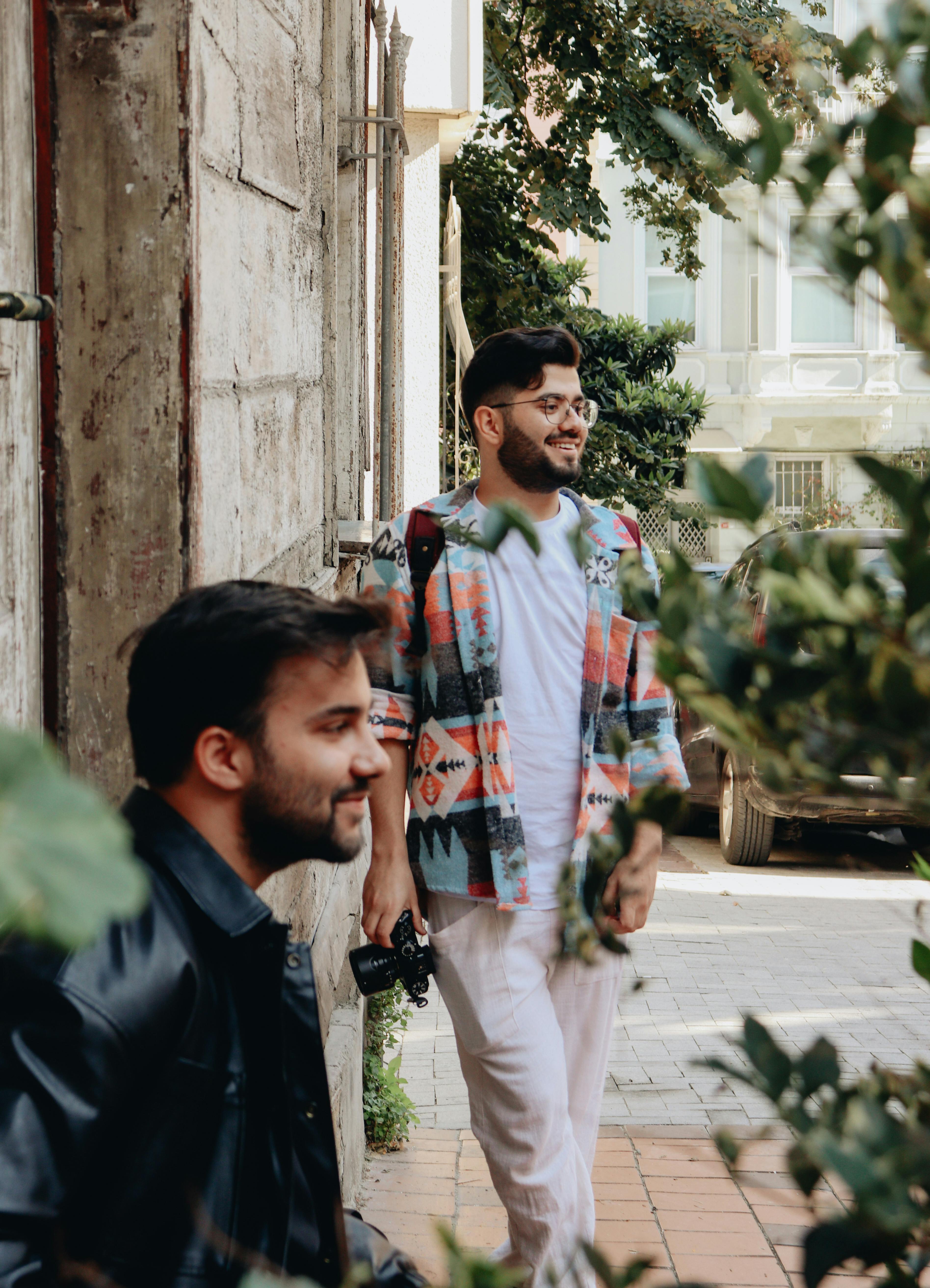 Two men on an urban street with one holding a camera, surrounded by greenery.