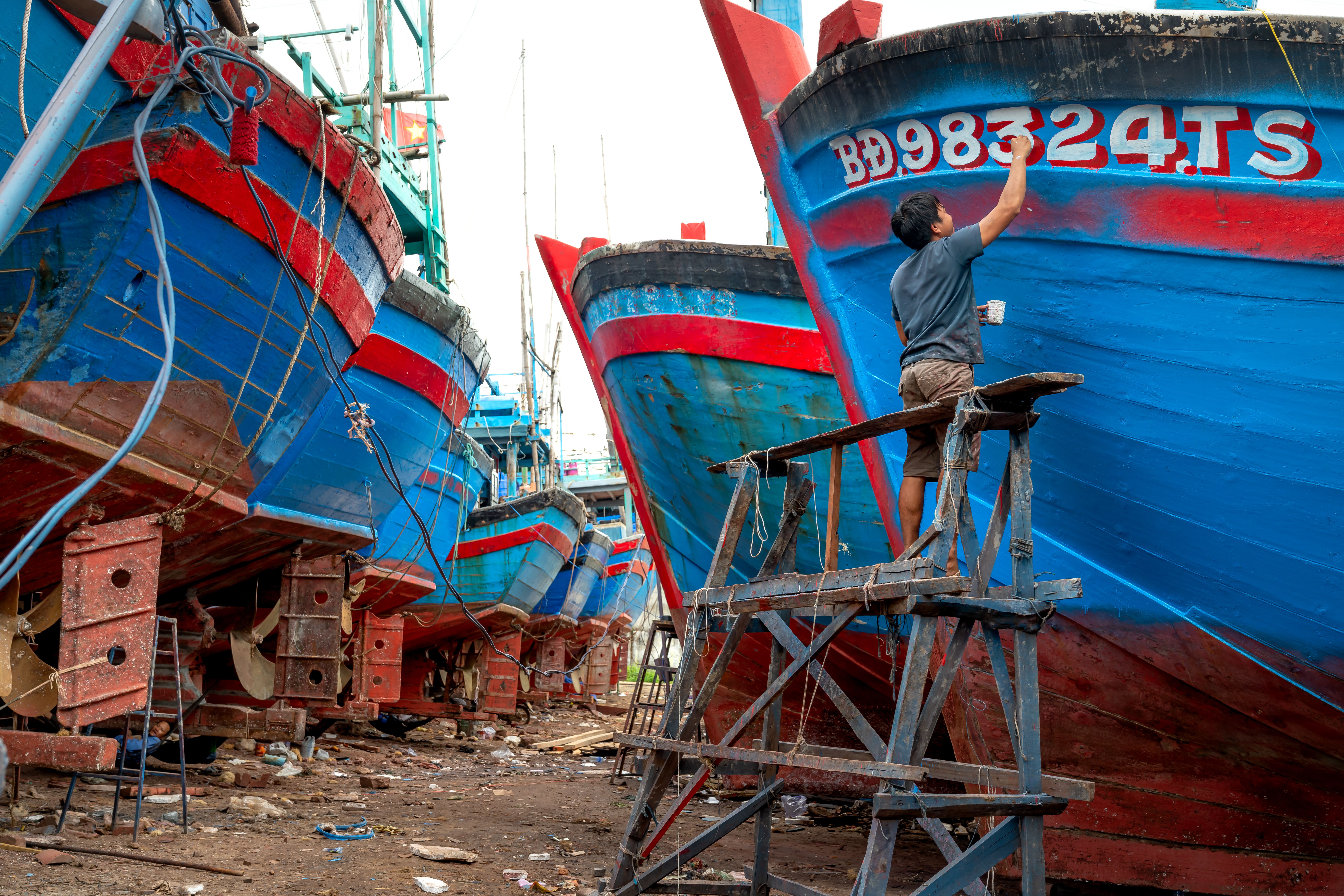 A man painting hulls of vibrant boats in an outdoor shipyard.