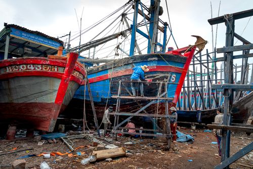 Free Laborers engaged in repairing fishing boats at an outdoor dockyard. Scaffolding surrounds the vessels. Stock Photo