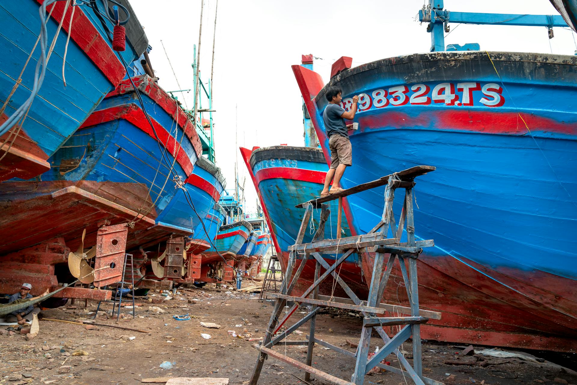 A man stands on scaffolding, painting a large wooden ship in a dry dock area.