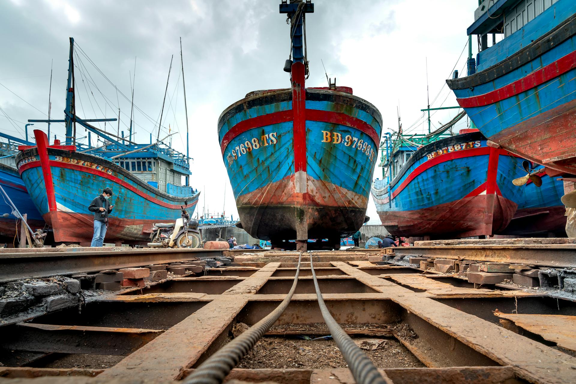 Vibrant fishing boats at an industrial shipyard with a worker and motorcycle nearby.
