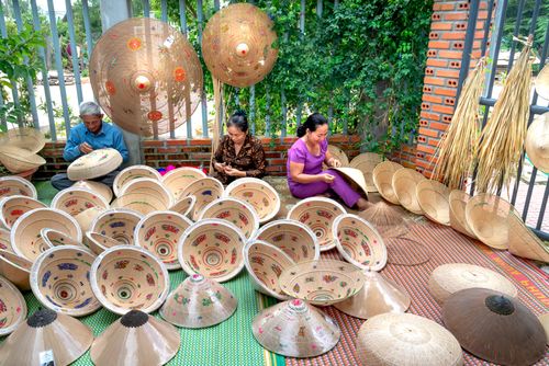 Free Artisans skillfully handcraft traditional Vietnamese conical hats outdoors surrounded by vibrant patterns. Stock Photo