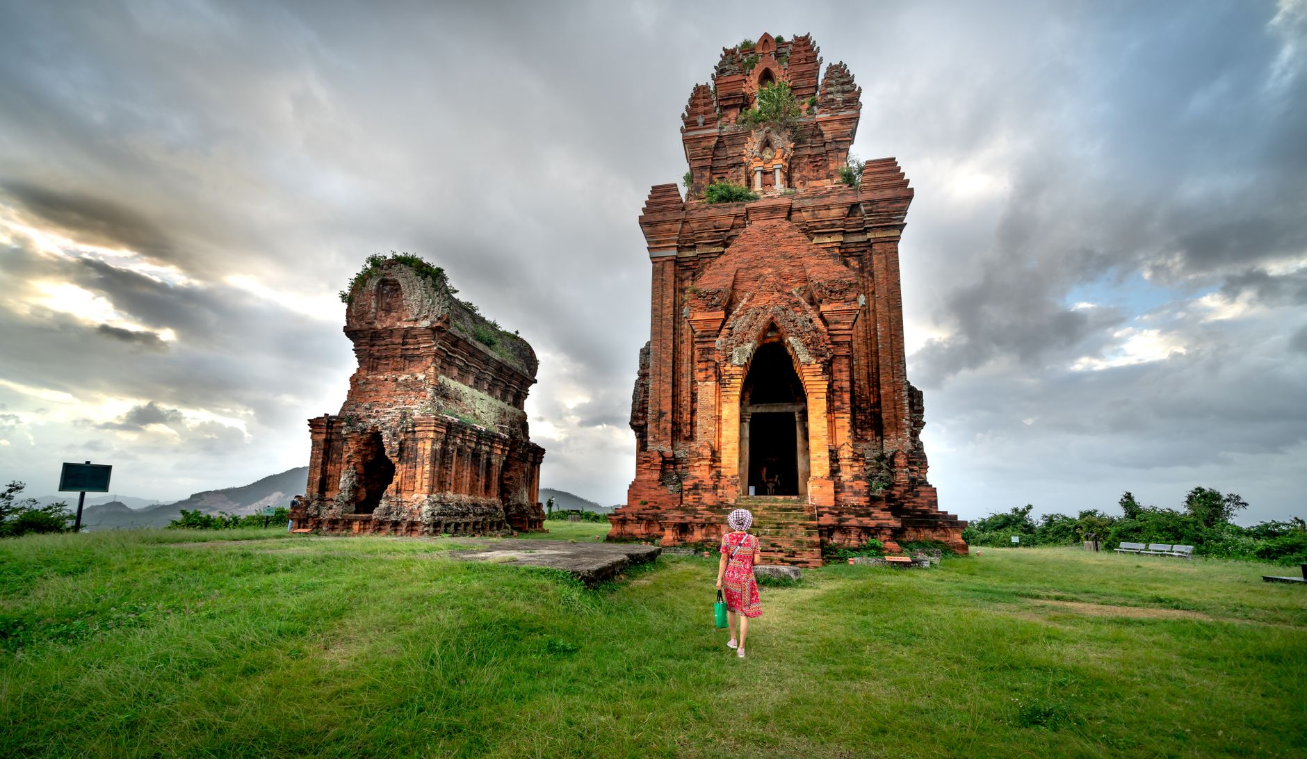 A woman walks towards ancient Cham temples under cloudy skies, surrounded by lush grass.