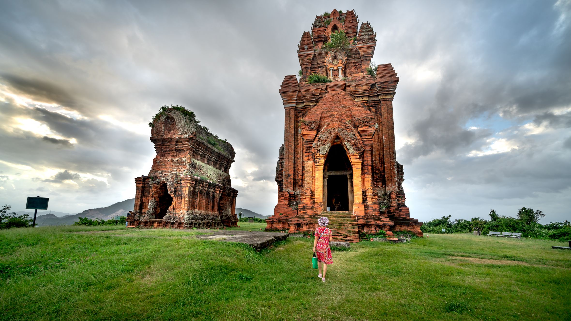 A woman walks towards ancient Cham temples under cloudy skies, surrounded by lush grass.