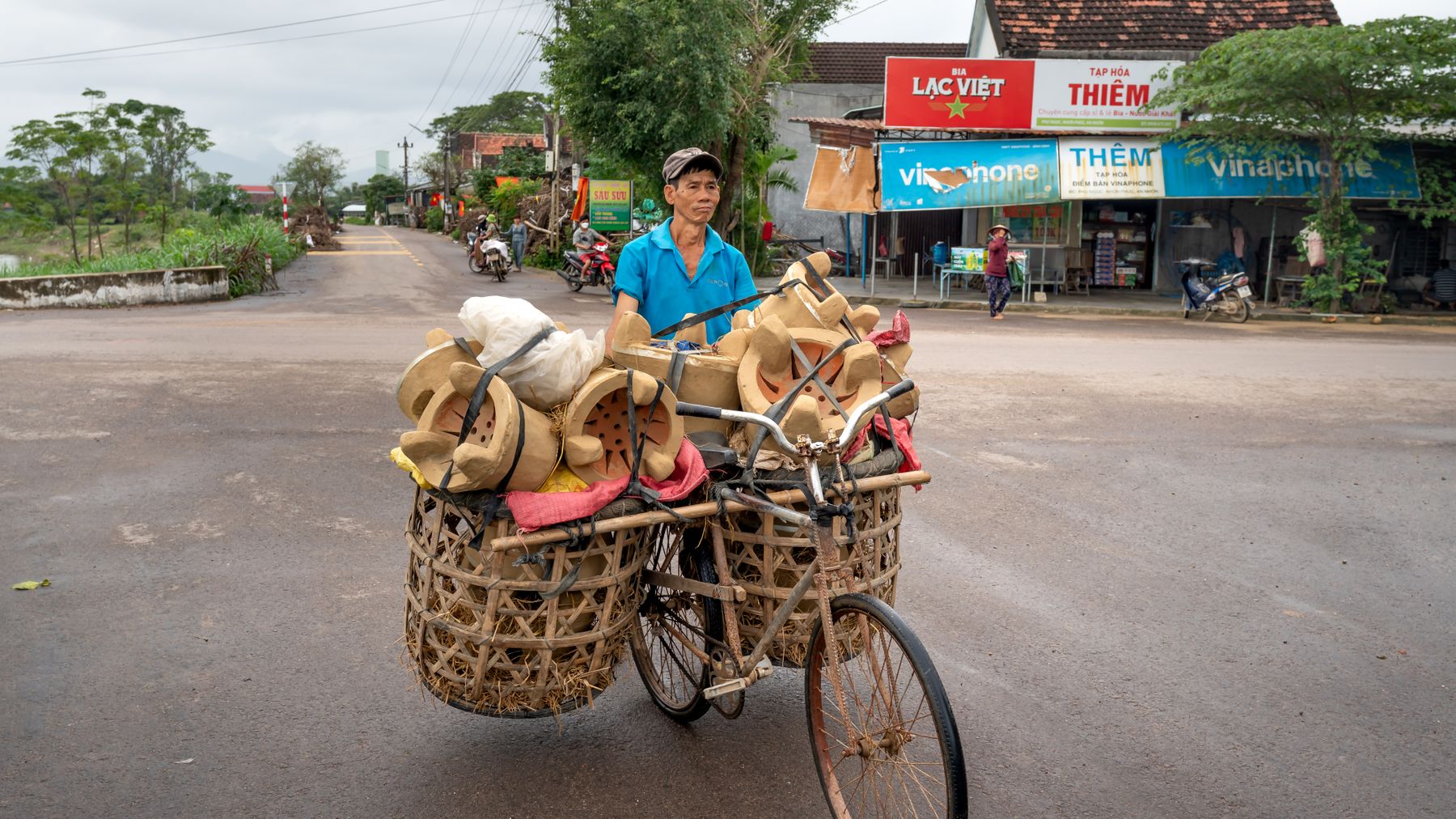 Asian man riding a bicycle transporting goods through a street in Vietnam. Townscape and shops visible in the background