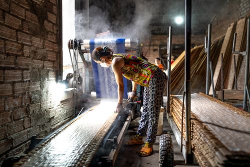 Free A female worker in vibrant clothing operates a machine in an industrial setting, with steam rising. Stock Photo