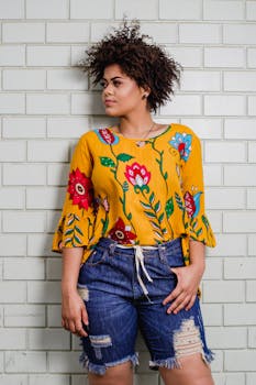 一位时尚的年轻女子身穿黄色花卉衬衫和牛仔短裤，倚着砖墙摆姿势