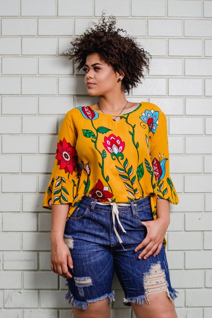Stylish young woman posing in a floral yellow blouse and denim shorts against a brick wall.