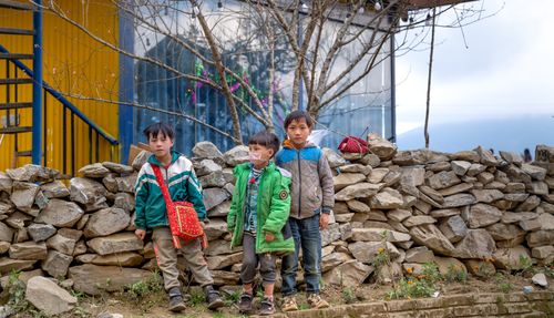 Free Three children in outerwear standing by a stone wall in a village setting. Stock Photo