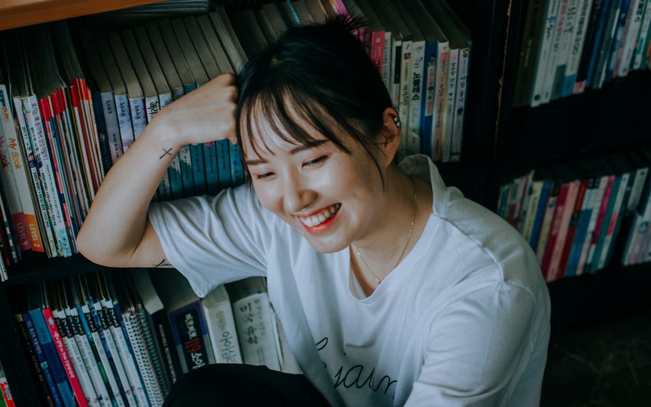 白いシャツを着た楽しい女性が、本でいっぱいの本棚のそばに座って微笑んでいます。