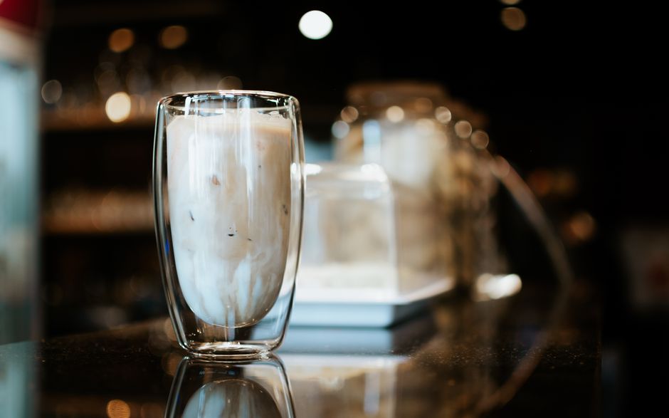 A glass of iced coffee on a reflective counter in a cozy café setting with warm lighting and bokeh effect.