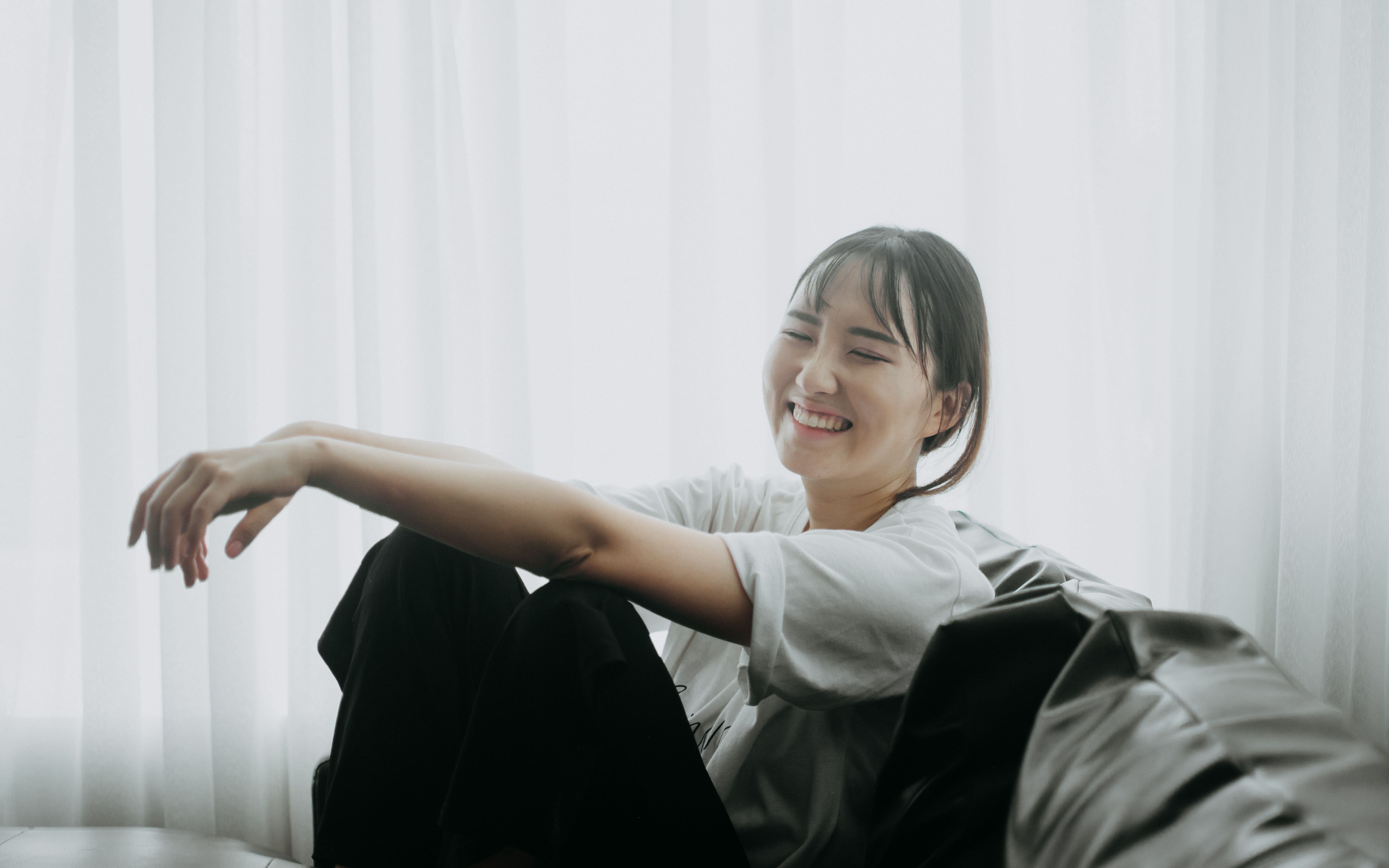 A cheerful Asian woman in a white t-shirt relaxes on a leather couch indoors.
